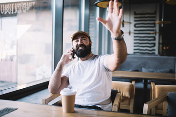 Smiling caucasian 40 years man in casual wear talking on mobile phone and raising hand for making order in cafe interior,cheerful prosperous mature male satisfied with positive smartphone conversation