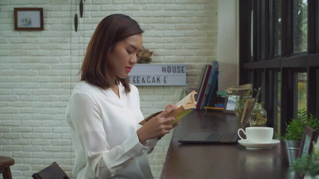 The asian woman is reading the book with laptop on the counter bar at home in the rainy day 