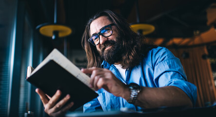 Smiling mature man in with long brunette hair spending free time on interesting literature hobby, positive caucasian male 40 years turn pages in book satisfied with leisure and self education in cafe