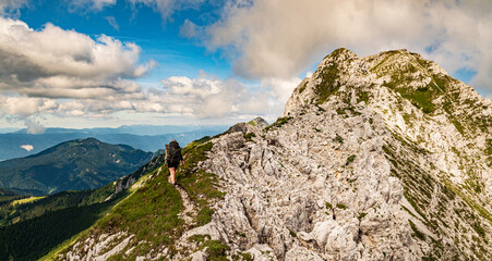 Young woman summer hiker with backpack climbing steep slopes mountains with valleys around. Female trekker in challenging terrain in Karavanke mountains, Slovenia and Karawanken, Carinthia, Austria
