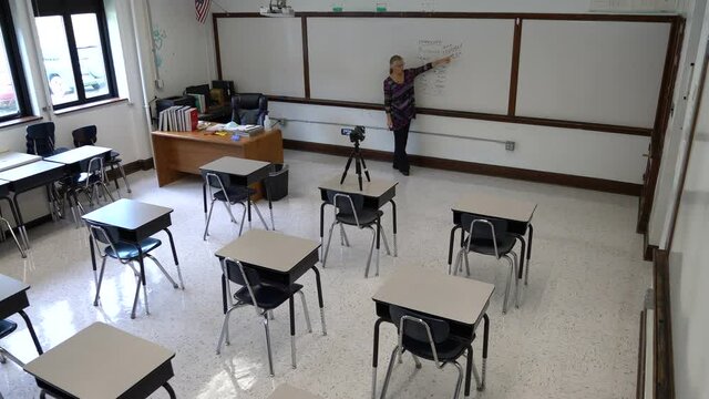 Wide angle view of smiling female english grammar teacher, writing on whiteboard in a school classroom with empty chairs.
