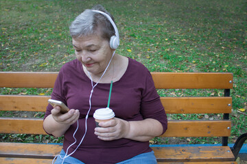 Elderly woman with headphones and cup of coffee sits on bench and talks on the phone with her...