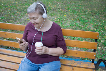 Elderly woman with headphones and cup of coffee sits on bench and talks on the phone with her family. The concept of  adaptation of older people in modern life. International Day of Older Persons