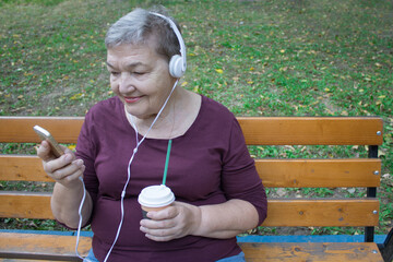 Elderly woman with headphones and cup of coffee sits on bench and talks on the phone with her...