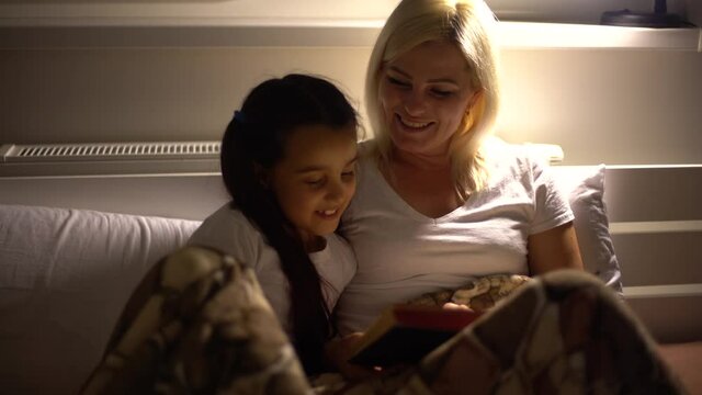 Happy Caucasian woman enjoying family time with her daughter at home together, smiling and talking in a bed, reading a book, with her daughter embracing, social