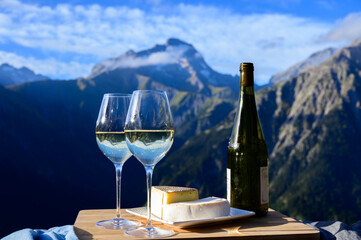 Tasty cheese and wine from Savoy region in France,  tomme and reblochon de savoie cheeses and glass of white wine served outdoor with Alpine mountains peaks on background - obrazy, fototapety, plakaty