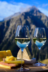 Tasty cheese and wine from Savoy region in France, beaufort, abondance, emmental, tomme and reblochon de savoie cheeses and glass of white wine served outdoor with Alpine mountains peaks on background