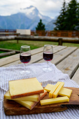 Cheese collection, French comte, beaufort or abondance cow milk cheese and glasses of red wine from Savoie served outdoor with Alps mountains peaks on background