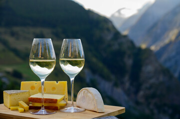 Tasty cheese and wine from Savoy region in France, beaufort, abondance, emmental, tomme and reblochon de savoie cheeses and glass of white wine served outdoor with Alpine mountains peaks on background