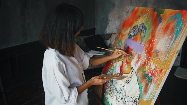 Young woman artist is painting a portrait on canvas in modern art style while working in studio