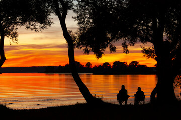 Horizontal landscape photography with black silhouettes of two unknown people sitting in the portable chairs on the lake shore under the silhouettes of trees with orange sunset on the background 
