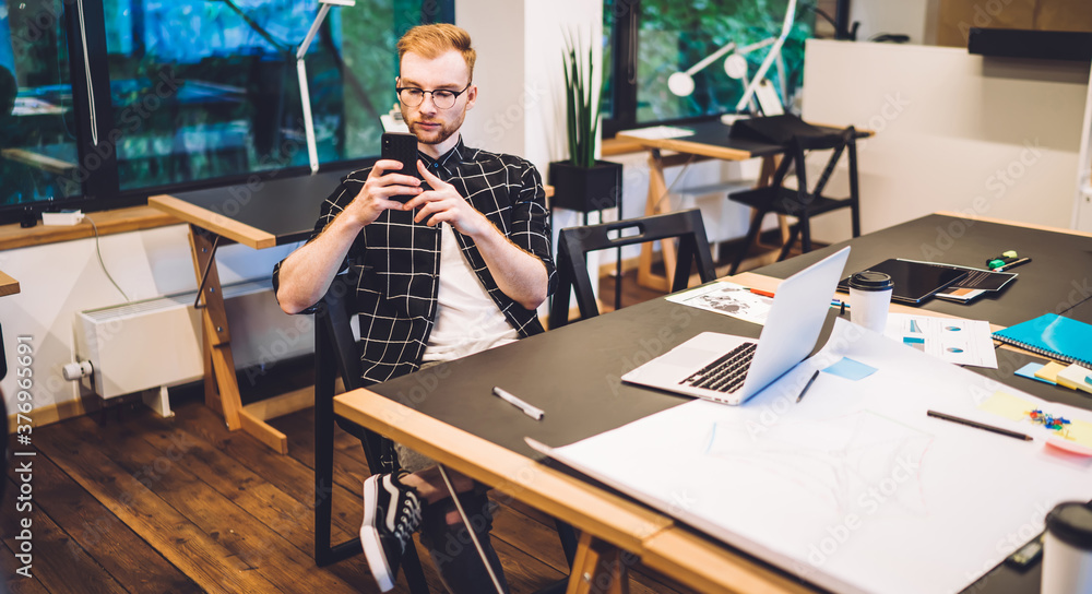Poster young hipster guy checking smartphone message during time for working with design project sitting at