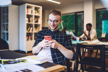 Young millennial hipster guy in optical spectales enjoying online chatting on smartphone device connecting to 4g indoors, Caucasian man checking content text for sharing mobile web publication