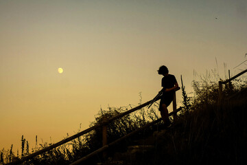 Silhouette of a young man coming down stairs on the mountain at dusk wih he moon in the sky