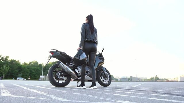 Back view of gorgeous sexy young stylish female biker with helmet in hands which coming up and getting on luxurious motorbike,looking at camera with nice smile