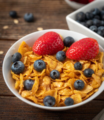 Corn flakes with blueberries and raspberries in white bowl in dark wooden desk.