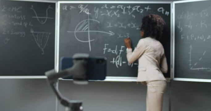 Mobile phone recording online video lesson at school. Lockdown isolated studying. African American woman teacher explaining math or physics formulas at class on blackboard. Pandemic education.