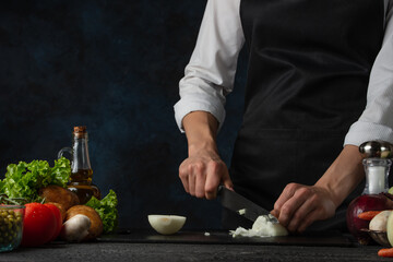 Chef cooking delicious food. Girl cut an onion. Girls hands with knife. Species on the table. Black background. Green salad, potatoes, tomatoes, onions and bottle with oil. Restaurants food.