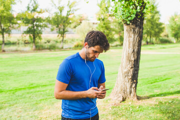 Attractive young man with a beard consults his smartphone dressed in sport clothes in the park.