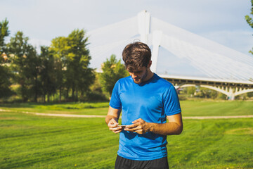 Attractive young man consults his smartphone dressed in sport clothes in the park.