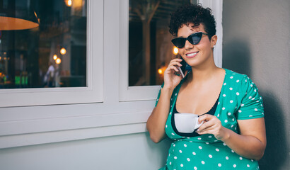 Smiling woman in sunglasses speaking on smartphone with cup in hand at table in outside cafe