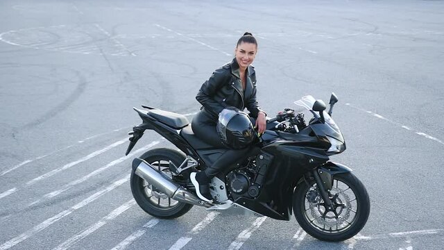 Portrait of splendid smiling satisfied young stylish female biker in leather clothes which posing on luxurious motorcycle on sportground in daytime