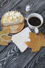 Cheesecake with white chocolate and a cup of coffee. The filling is visible on the cut of the dessert. Nearby is a medical mask, antiseptic and dried maple leaves. On pine planks.