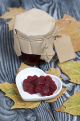 A jar of cherry jam with a label for an inscription and a saucer of cherry marmalade. Dried maple leaves all around. On pine planks painted black and white.