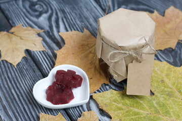 A jar of cherry jam with a label for an inscription and a saucer of cherry marmalade. Dried maple leaves all around. On pine planks painted black and white.
