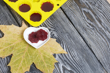Saucer with cherry marmalade. A silicone mold with marmalade is visible. Dried maple leaves all around. On pine planks painted black and white.