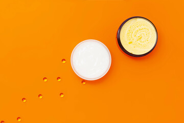 Two jars with different creams on an orange background next to which are capsules with vitamin E.