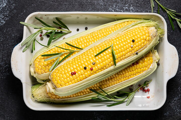 Raw corn cobs with spices and herbs ready to grill in ceramic dish on black rustic background top view. Healthy food concept.