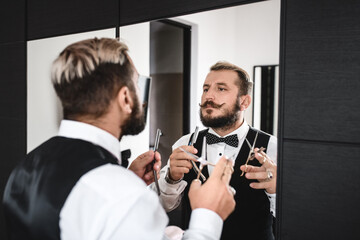 A stylish man with a luxurious mustache and beard holds blades and scissors in his hands. He stands in front of the mirror. Barber