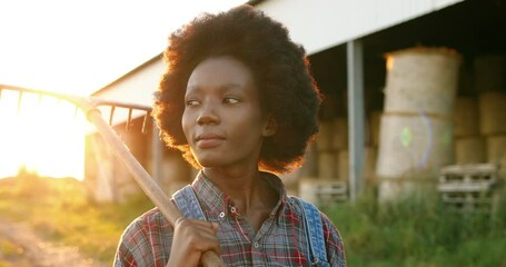 African American woman with curly hair walking in field early in morning with pitchfork. Farming lifestyle. female farmer holding fork and going to work outdoor. Girl in village.