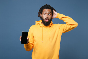 Shocked young african american man guy in yellow hoodie isolated on blue background studio. People lifestyle concept. Mock up copy space. Hold mobile phone with blank empty screen put hand on head.