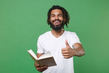 Cheerful funny young african american man guy with dreadlocks 20s wearing white casual t-shirt posing holding in hand reading book showing thumb up isolated on green color background studio portrait.
