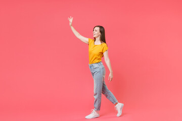 Fototapeta na wymiar Full length portrait side view of cheerful young brunette woman 20s wearing yellow casual t-shirt posing waving and greeting with hand as notices someone isolated on pink color wall background studio.