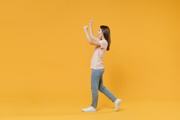 Fototapeta na wymiar Full length side view portrait of happy joyful young woman wearing pastel pink casual t-shirt posing clenching fists doing winner gesture rising hands isolated on yellow color wall background studio.