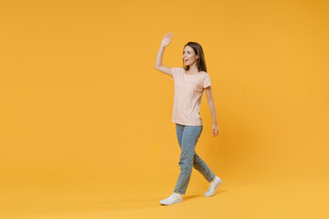Fototapeta na wymiar Full length portrait of cheerful funny young woman 20s in pastel pink casual t-shirt posing waving and greeting with hand as notices someone looking aside isolated on yellow color background studio.