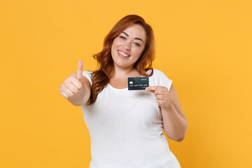 Smiling young redhead plus size body positive female woman girl 20s in white blank casual t-shirt posing hold credit bank card showing thumb up isolated on yellow color background studio portrait.