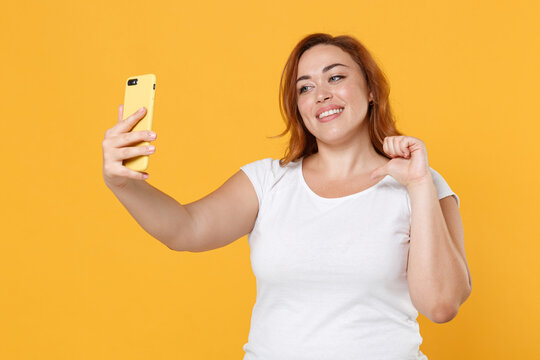 Smiling young redhead plus size body positive female woman girl in white casual t-shirt doing selfie shot on mobile phone pointing thumb on herself isolated on yellow color background studio portrait.