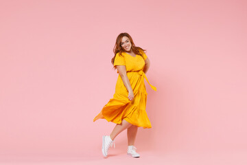 Full length portrait of smiling beautiful charming young redhead plus size body positive female woman girl 20s in yellow dress posing looking camera isolated on pastel pink color background studio. - obrazy, fototapety, plakaty