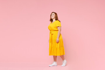 Full length portrait of smiling attractive beautiful young redhead plus size body positive female woman girl 20s in yellow dress posing looking aside isolated on pastel pink color background studio.