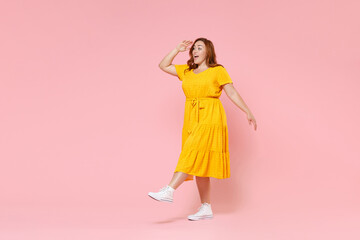 Full length portrait excited young redhead plus size body positive female woman 20s in yellow dress holding hand at forehead looking far away distance isolated on pastel pink color background studio.