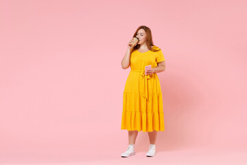 Full length portrait pretty young redhead plus size body positive female woman 20s in yellow dress hold paper cup coffee tea mobile phone looking aside isolated on pastel pink color background studio.