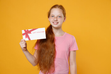 Smiling little ginger redhead kid girl 12-13 years old wearing pink casual t-shirt posing hold gift certificate looking camera isolated on bright yellow color wall background children studio portrait.