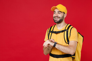 Delivery man in yellow cap t-shirt uniform thermal bag backpack with food hand smartwatch isolated on red background studio Guy male employee work as courier Service coronavirus covid-19 virus concept