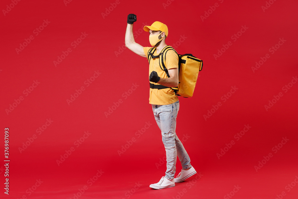 Wall mural Full length delivery employee man in yellow cap face mask gloves t-shirt uniform thermal food bag backpack work courier service during quarantine coronavirus covid-19 virus isolated on red background.