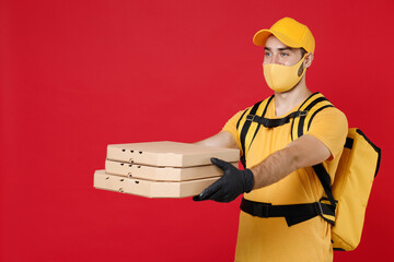 Delivery employee man in yellow cap face mask gloves t-shirt thermal bag backpack with food hold pizza in cardboard flatbox work courier service quarantine covid-19 virus isolated on red background.