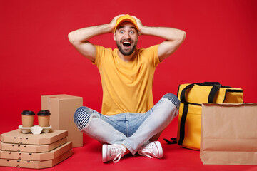 Delivery employee man guy male in yellow cap t-shirt sit at thermal takeaway food bag backpack pizza boxes cups work courier service during quarantine coronavirus covid-19 isolated on red background.